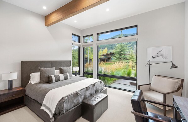 A modern bedroom with a large bed, a seating area, and large windows offering a view of a forest and a building. - The Cabins At Snake River Sporting Club (Jackson)