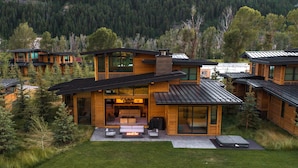Une maison moderne à plusieurs étages, dotée de grandes fenêtres, d’un patio avec des meubles d’extérieur et offrant une vue sur une chaîne de montagnes boisée.