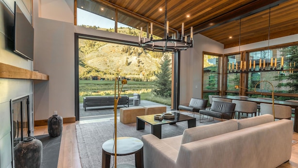 Un salon moderne avec un grand canapé, une table basse et une cheminée. De grandes fenêtres offrent une vue sur un jardin et des collines.