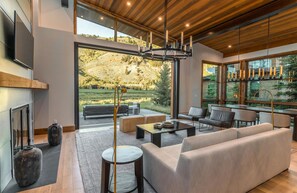 A modern living room with a large sofa, coffee table, and a fireplace. There are large windows offering a view of a garden and hills. - The Cabins At Snake River Sporting Club (Jackson)
