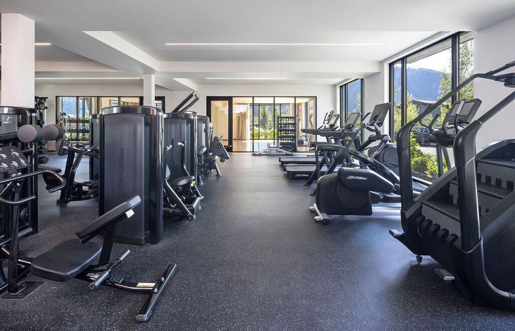 A modern gym with cardio machines, weight stations, and a large window offering a view of the outdoors.