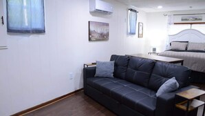 A room with a black leather sofa, a bed with a white headboard, a small table, and a wall-mounted air conditioner. - The Center Street Studio - Sleeps 4 (Searcy)