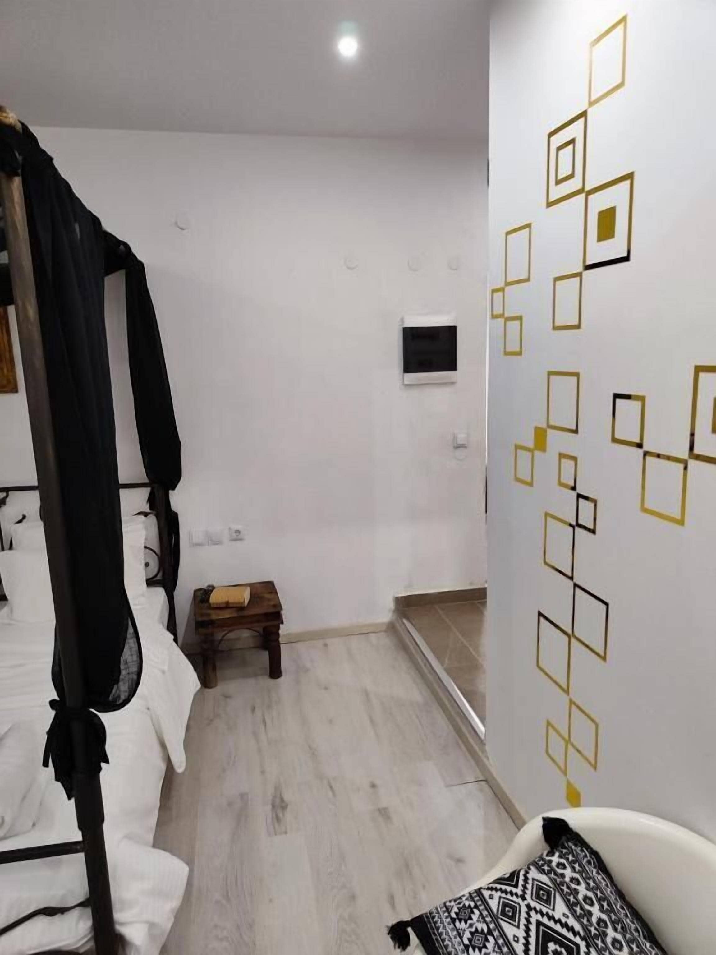 Een slaapkamer met een bed, een klein houten krukje, een witte stoel en een muur met geometrische patronen.