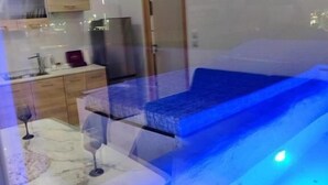 A modern room with a glass partition, a bed, a small table with a wine glass, and a kitchenette area. - Anchor Suites (Thessaloniki)