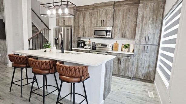 A modern kitchen with a large island, bar stools, wooden cabinets, stainless steel appliances, and a staircase leading to another level.