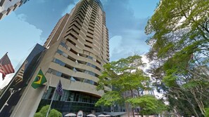 A tall, modern building with a curved facade, multiple flags, and surrounding greenery. - Suíte na Berrini by Conviva (Sao Paulo)