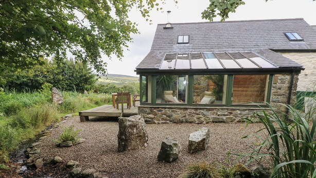 Una casa in pietra con veranda in vetro, un patio in legno e un paesaggio naturale con rocce e piante.