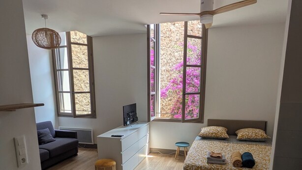 A modern, compact living space with a kitchenette, a dining area, and a bedroom with a large window. - Apartment Sète centre-ville (Sète)
