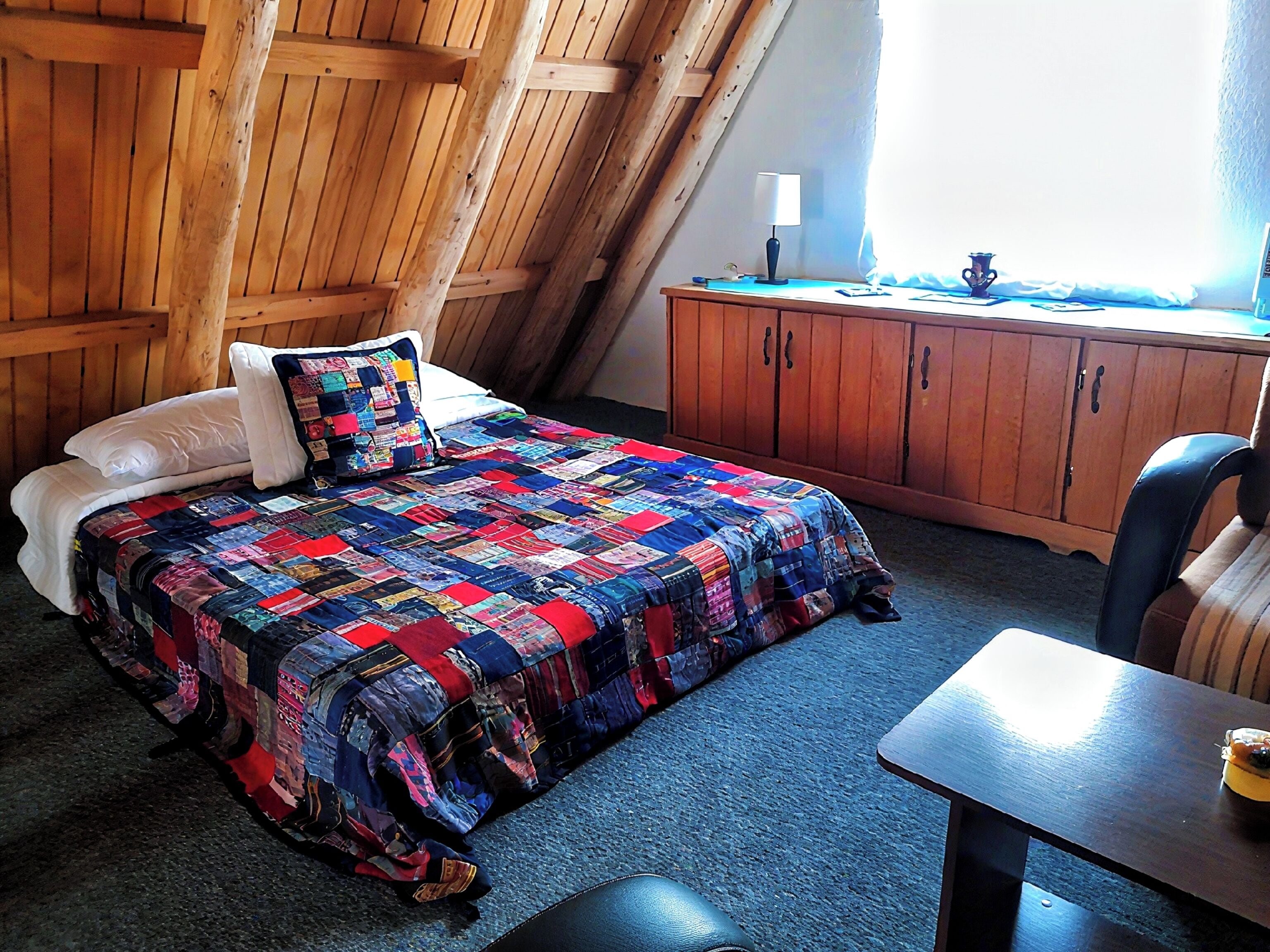 Une chambre avec un plafond en pente en bois, un lit recouvert d’une courtepointe colorée, une armoire en bois et une table sur laquelle se trouve une tasse.