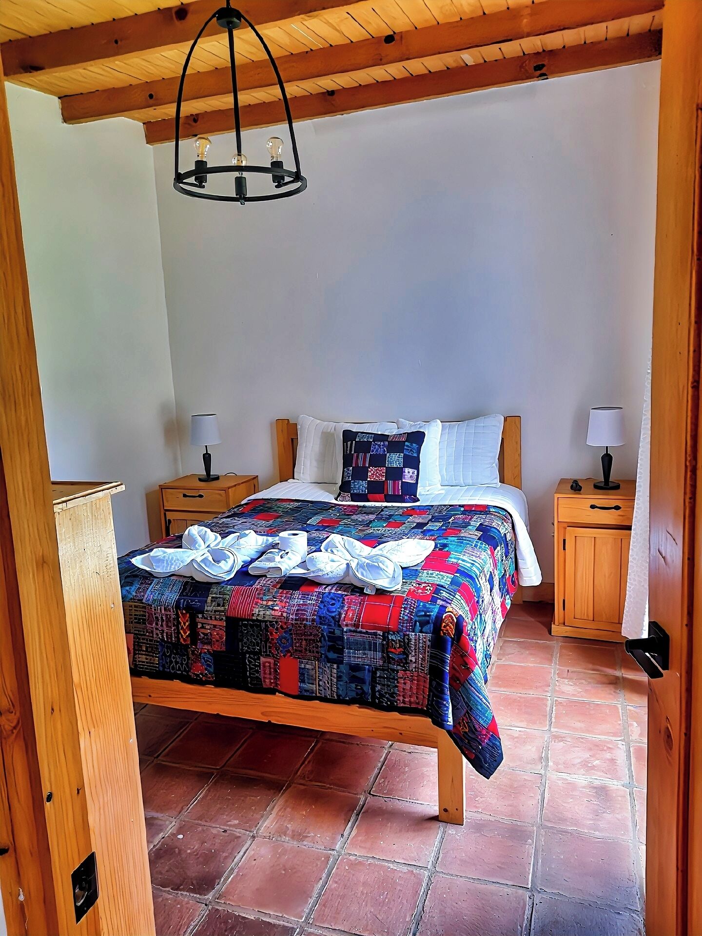 Une chambre à coucher avec un lit en bois, une courtepointe aux couleurs vives, deux tables de chevet avec des lampes et un plafond en bois.