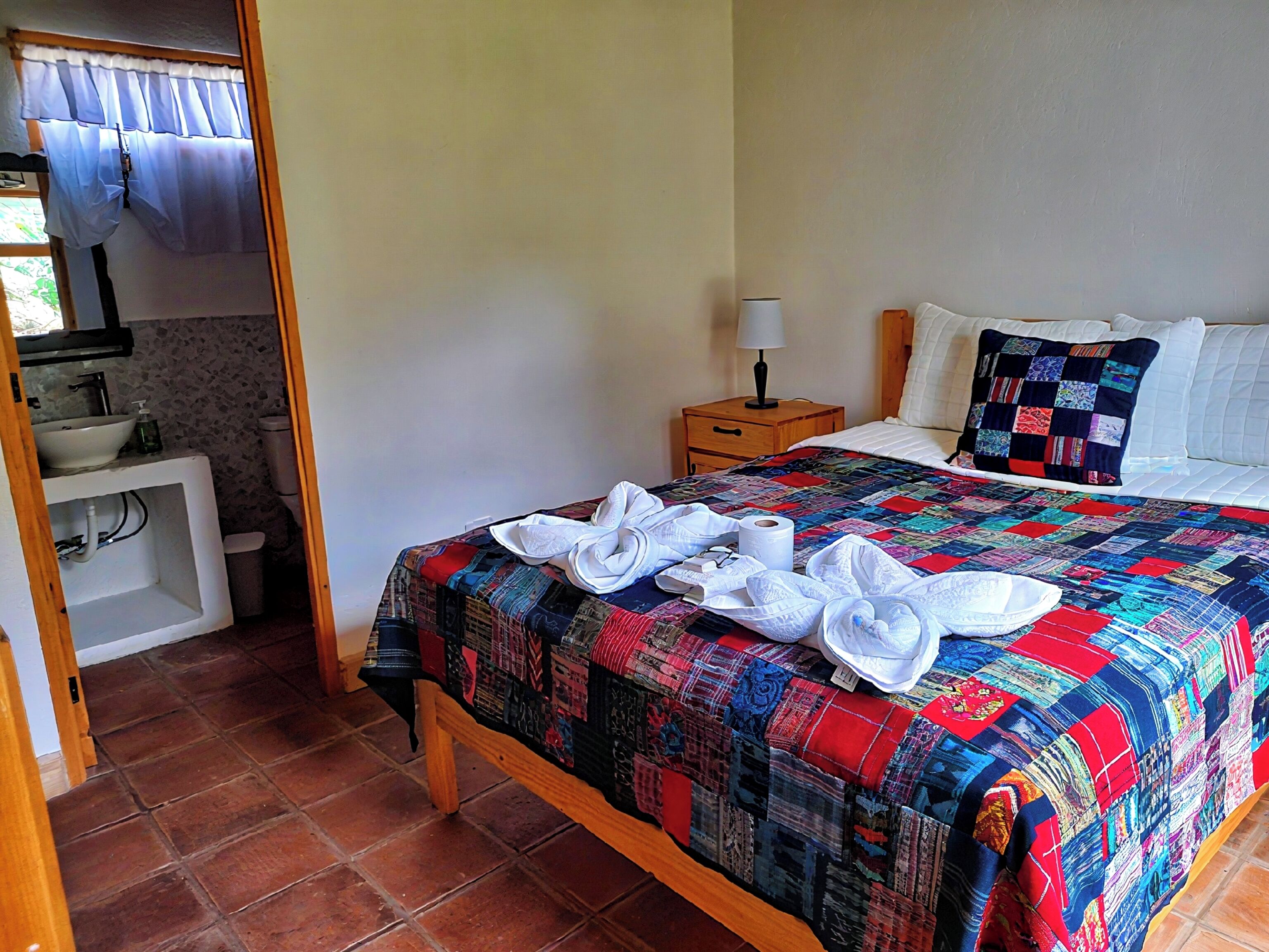 Une chambre d’hôtel comprenant un lit, une table de chevet avec une lampe, une salle de bain avec un lavabo et des toilettes, et une fenêtre avec des rideaux.
