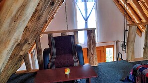 Une pièce mansardée chaleureuse avec un plafond en bois, une grande fenêtre avec des rideaux, une chaise à rayures, une petite table sur laquelle se trouve un fruit, et une couverture colorée à rayures posée sur un banc.