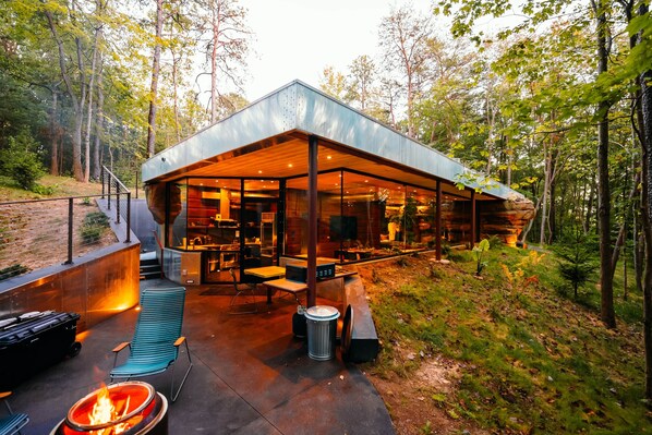 Un espacio moderno para comer al aire libre con una chimenea, asientos y vistas al bosque.
