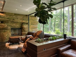A modern interior with a stone wall, leather furniture, a fireplace, and a large window with a forest view.