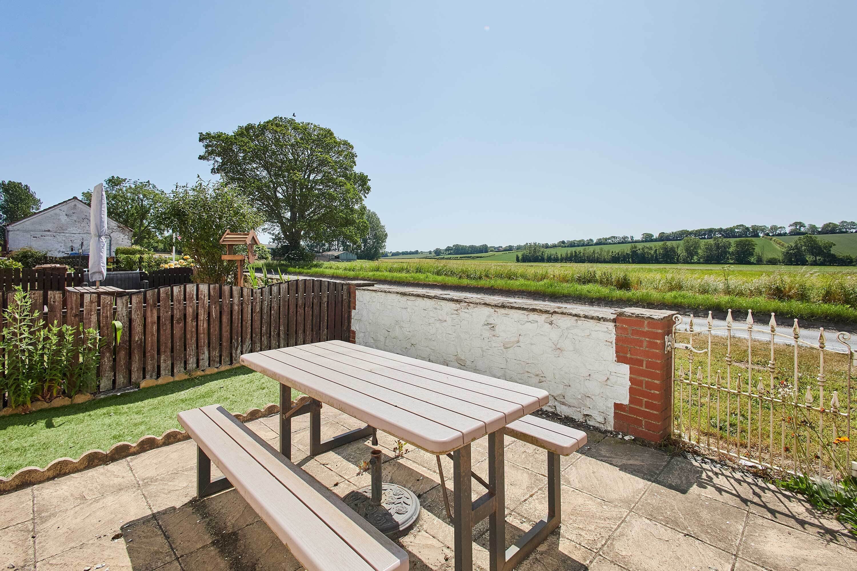 A patio with a wooden table and benches, a brick wall, and a view of a green field.
