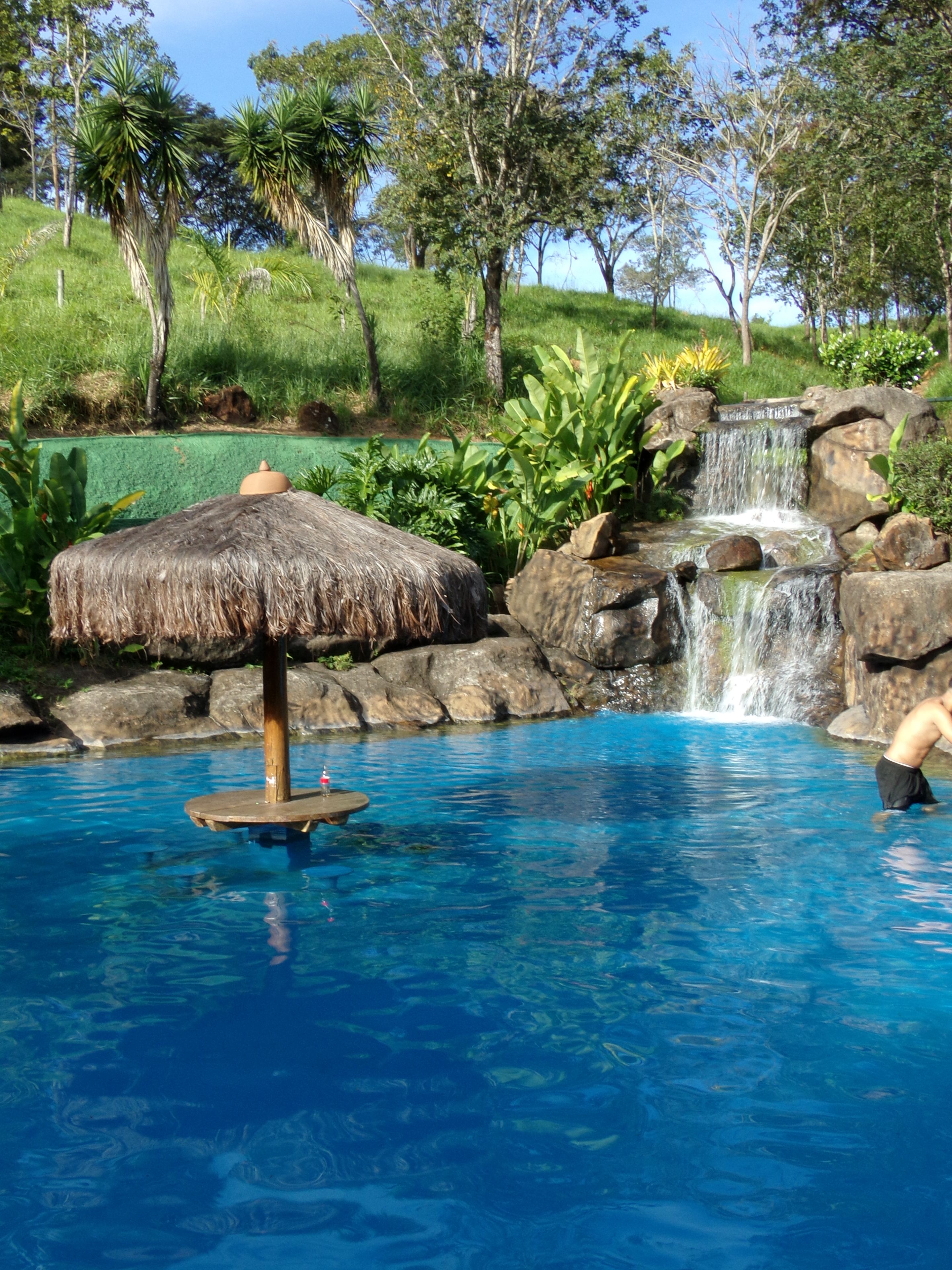 A clear pool with a thatched umbrella, a waterfall, and lush greenery in the background.