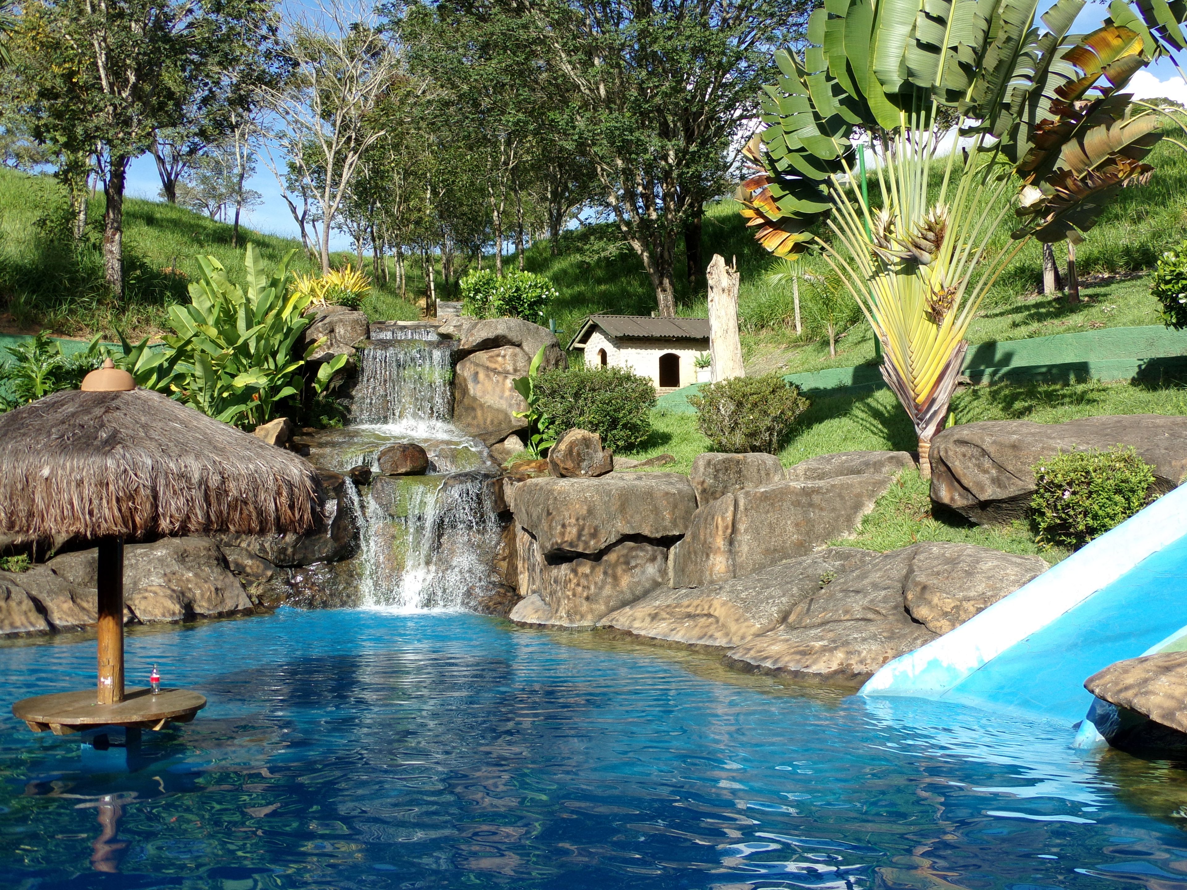 Piscina transparente com cascata, rodeada por pedras e vegetação.