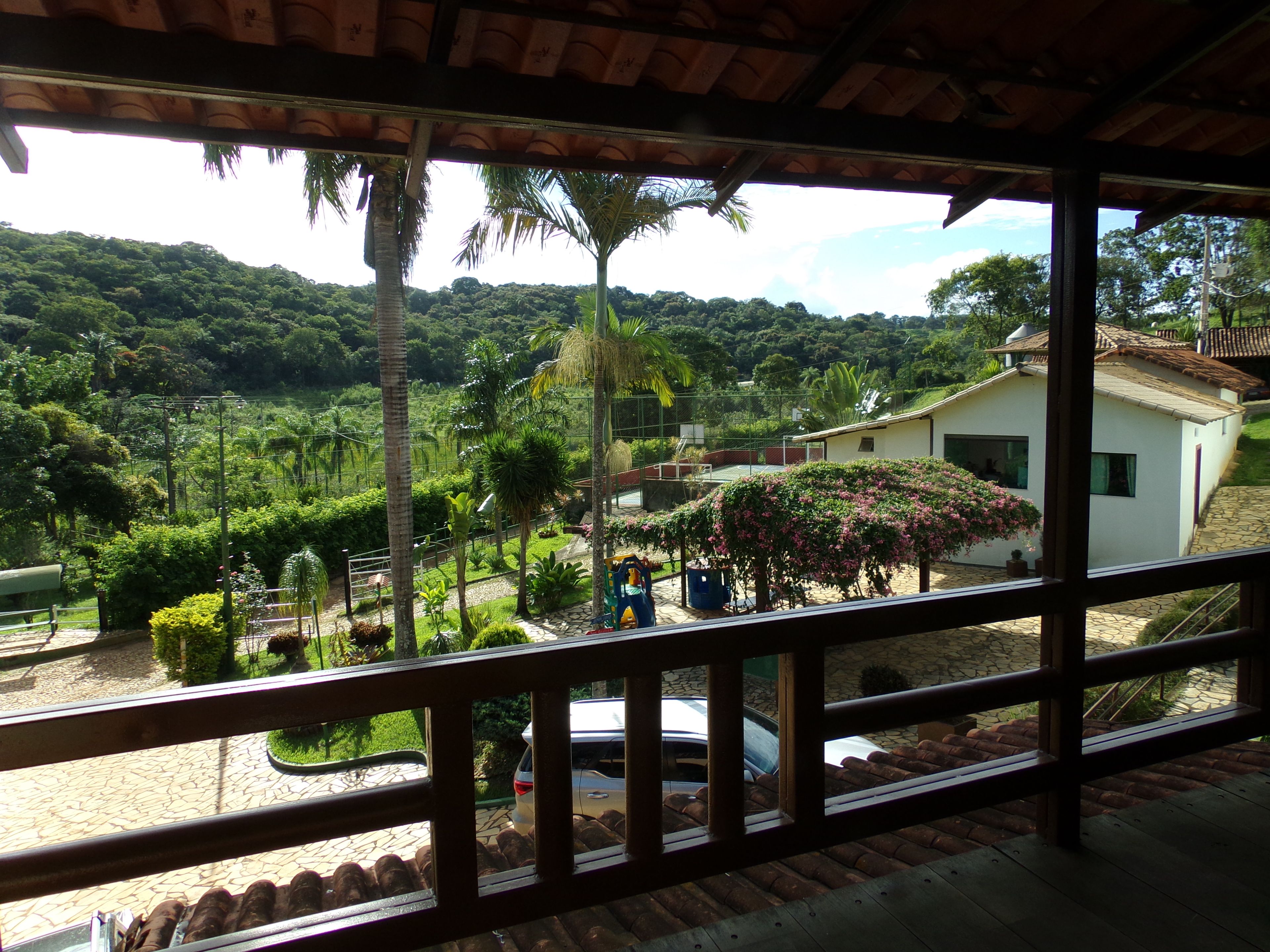 Vista de uma varanda coberta com piso de pedra, com vista para um jardim exuberante com palmeiras e um edifício branco com telhado ladrilhado.