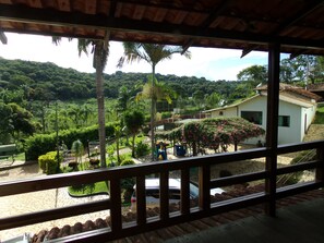 Vista desde un balcón cubierto con suelo de piedra, sobre un jardín exuberante con palmeras y un edificio blanco con techo de tejas.