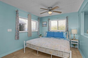 Une chambre avec un lit simple, un ventilateur de plafond, deux fenêtres avec des rideaux, une table de chevet avec une lampe et une petite plante.