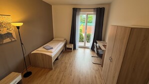 A small, well-lit room with a bed, a desk, a chair, a wardrobe, and a floor lamp.