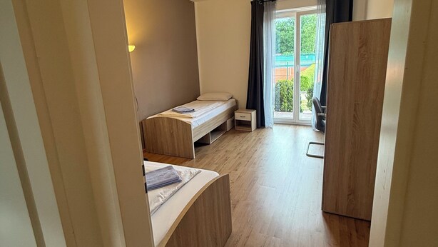 A compact room with a bed, a small desk, and a wardrobe. There is a window with curtains and a view of greenery outside.