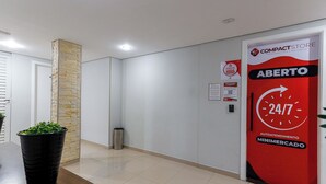 Un vestíbulo de hotel con mostrador de recepción, un muro decorativo de piedra, un cubo de basura y una puerta con un letrero rojo que indica disponibilidad las 24 horas.