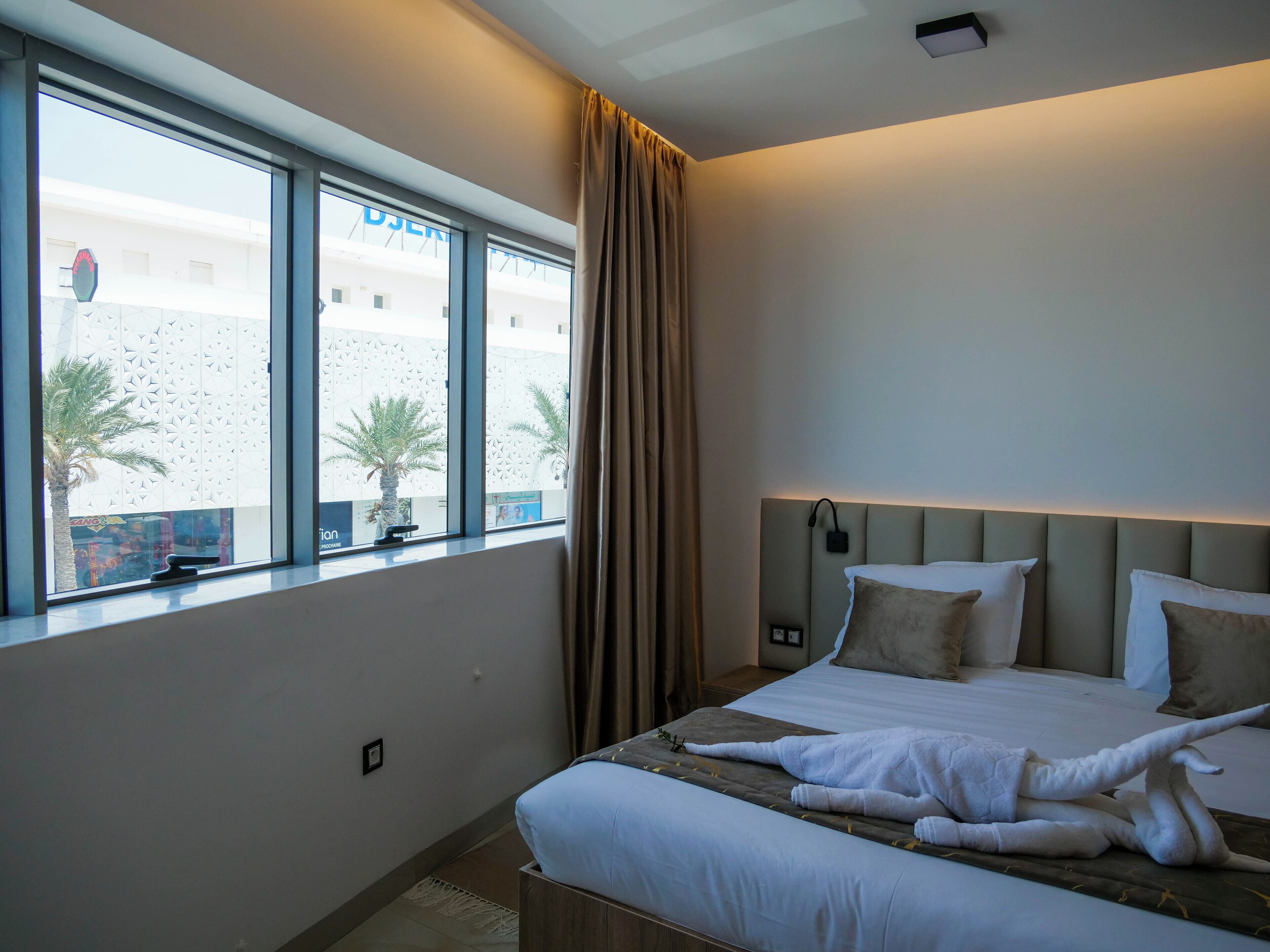 Hotelkamer met een groot bed, hoofdeinde met ingebouwde verlichting, raam met uitzicht op een gebouw en palmbomen, en een gordijn voor het raam.