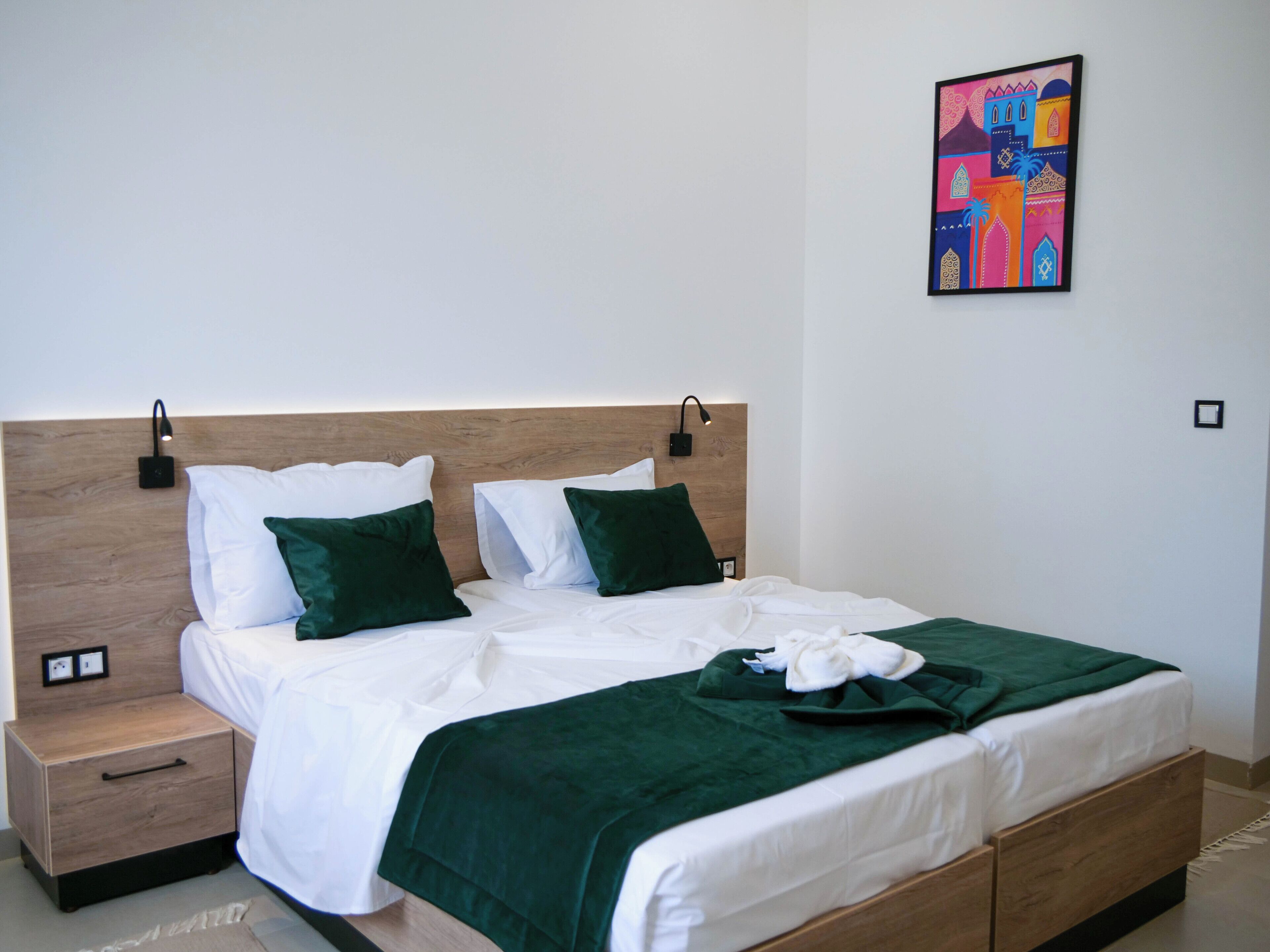 Un letto rifatto con lenzuola bianche e cuscini verdi, una testiera in legno, un comodino con un cassetto e un quadro colorato appeso alla parete.