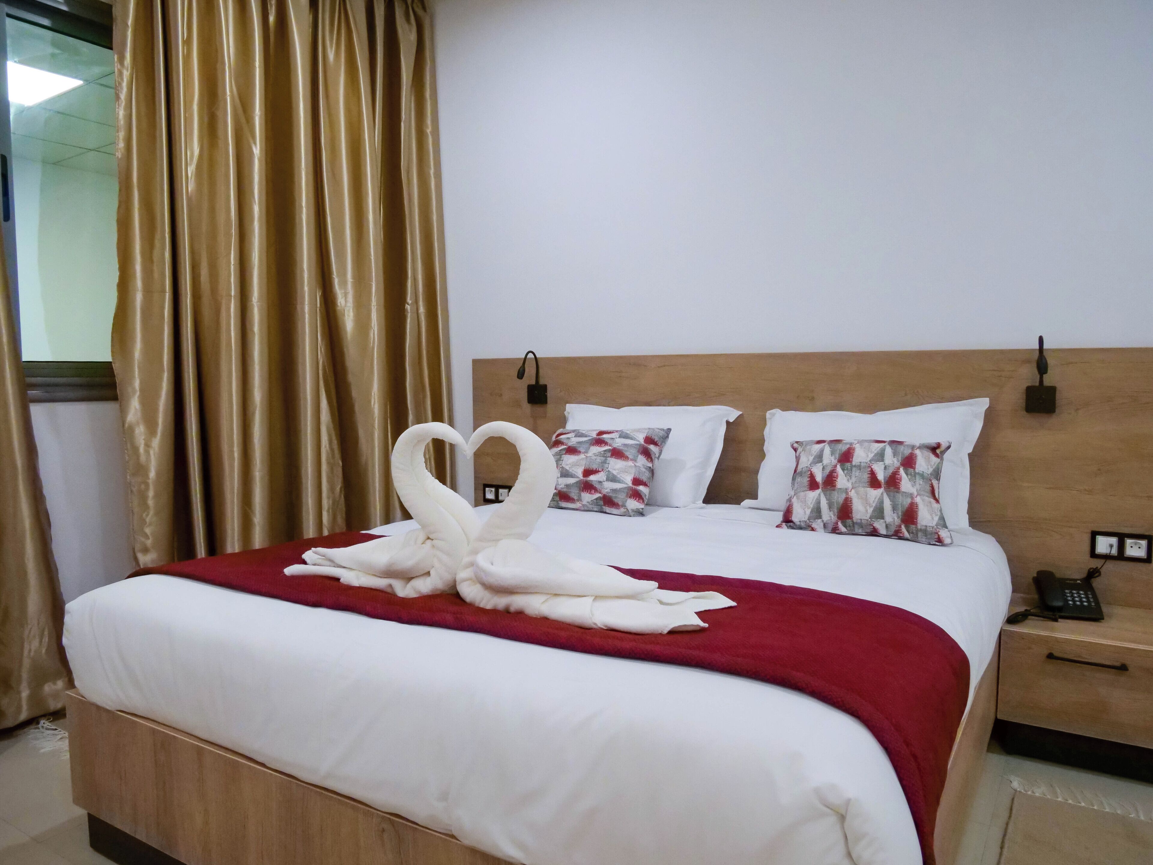 Ein Hotelzimmer mit einem ordentlich bezogenen Bett, einem hölzernen Kopfteil, einem Nachttisch mit Telefon und einem Fenster mit Vorhängen.