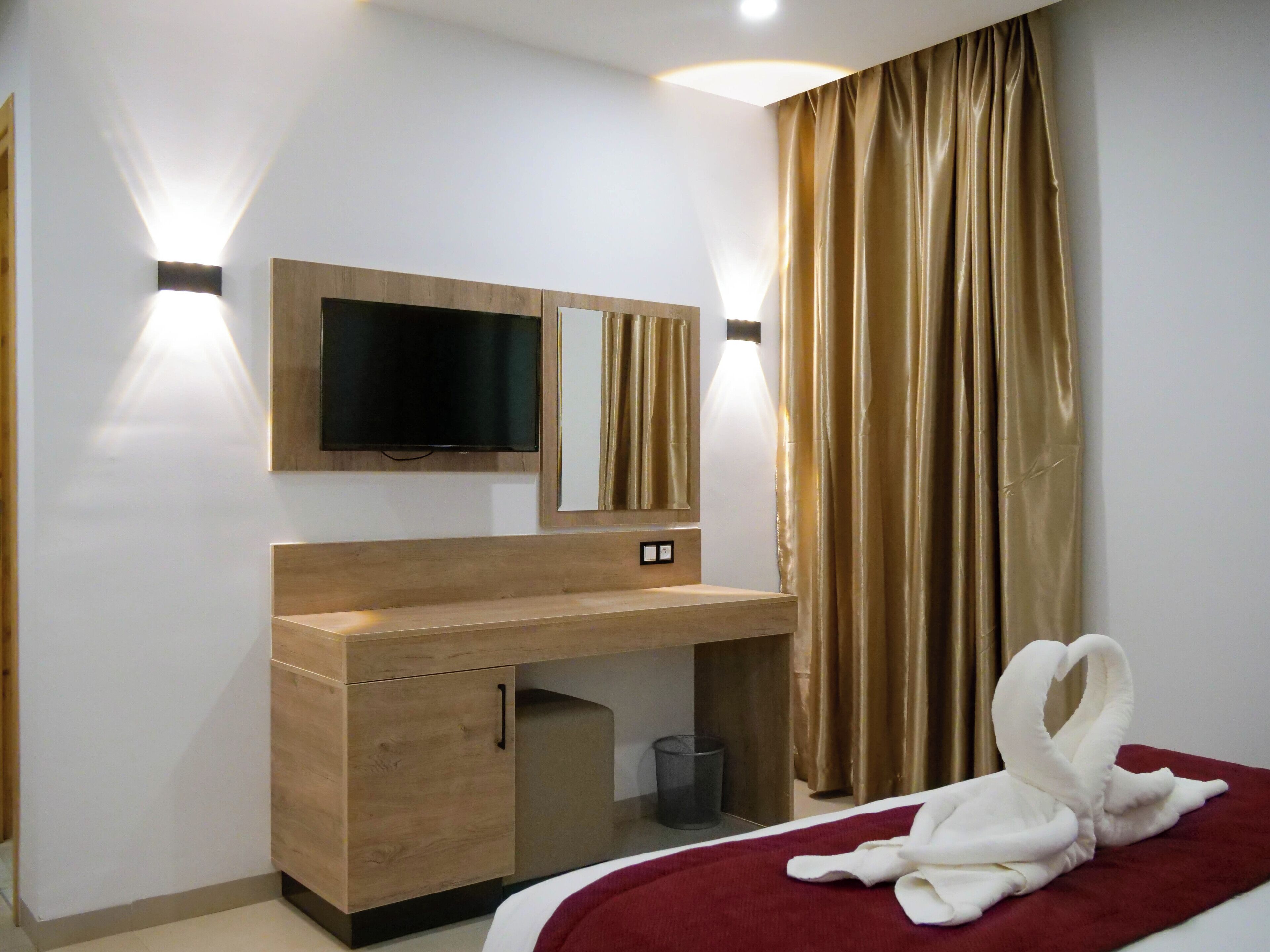 A hotel room with a wooden desk, a flat-screen TV mounted above it, a bed with a red blanket, and a golden curtain.