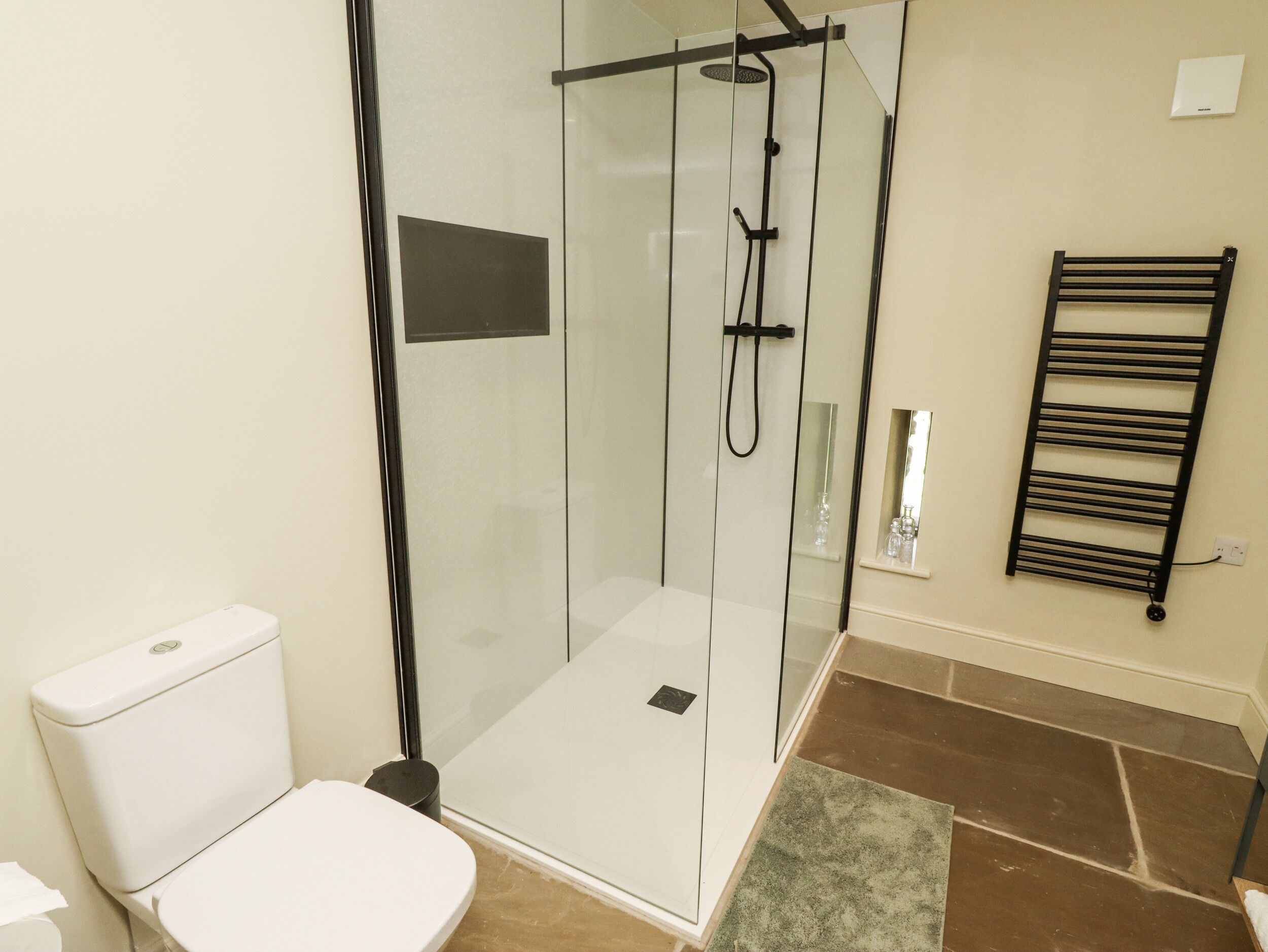 Un baño moderno con ducha de vidrio, inodoro blanco, toallero negro y suelo de baldosas.