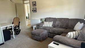 A living room with a sofa, ottoman, and a small table. There is a chair, a radiator, and a door leading to another room. - Cosy Chichester Apartment (Chichester)