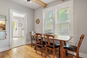 A dining area with a wooden table and chairs, a window with blinds, and a kitchen visible through an open door.
