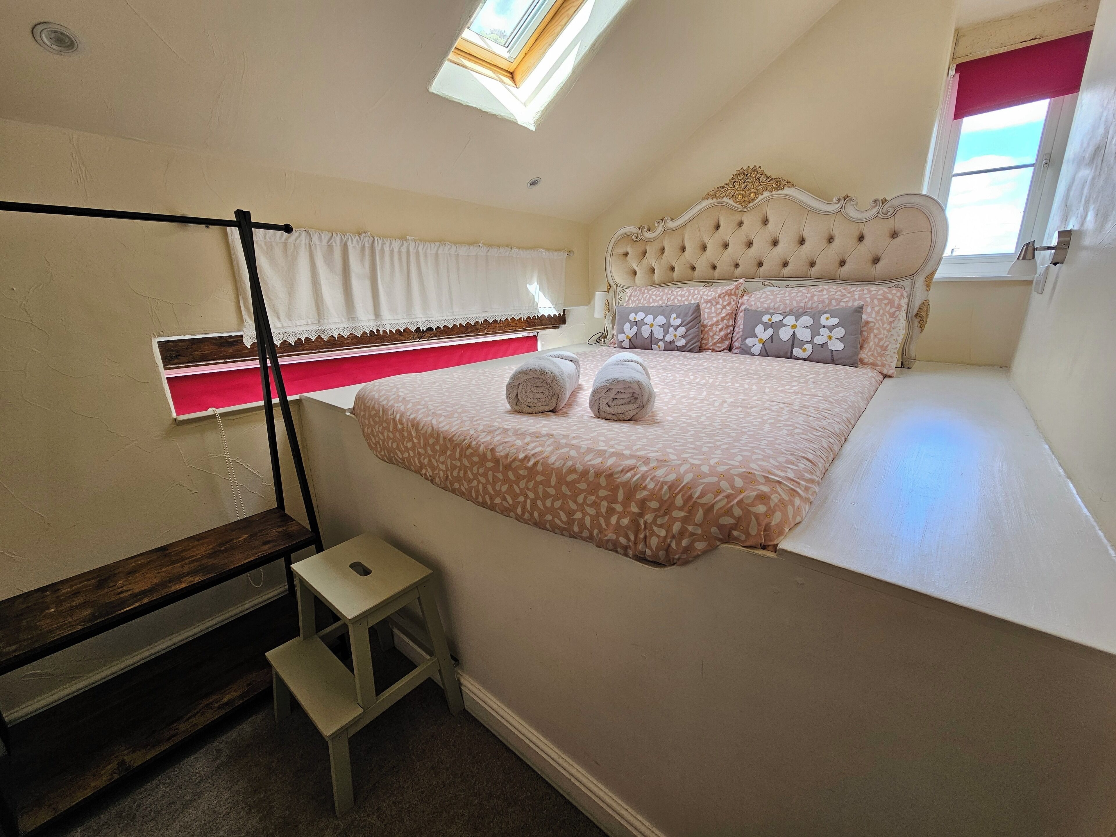 Un dormitorio con una cama, un taburete pequeño, una ventana con cortina y un tragaluz.