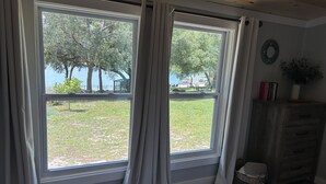 Ein Schlafzimmer mit großem Fenster, einem Bett, einem Schminktisch und Blick auf einen Rasen und Bäume.