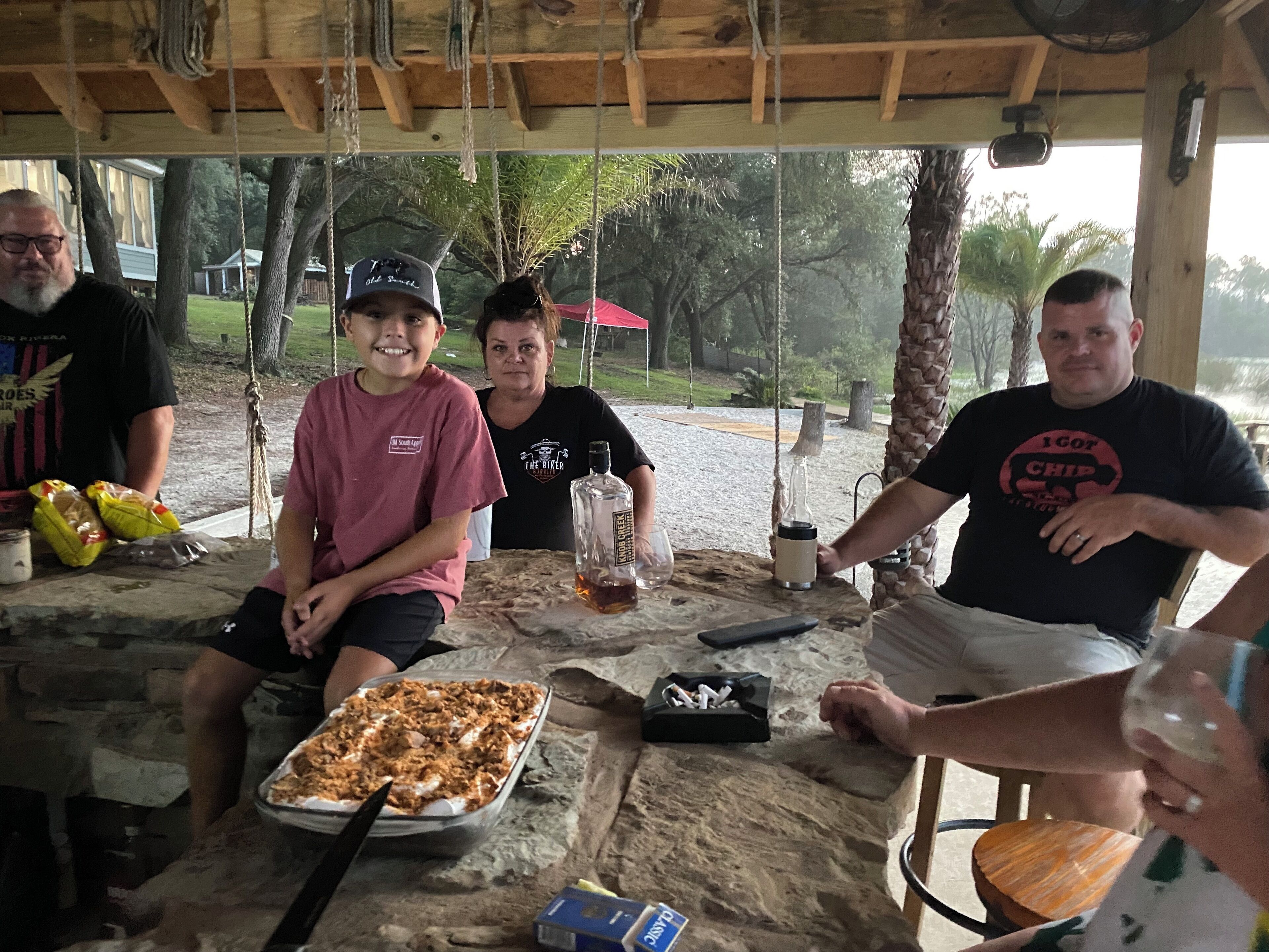 Un grupo de personas sentadas en una mesa al aire libre, con una bandeja de comida, una botella y un paquete de cigarrillos.