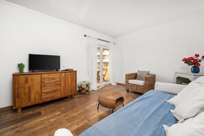 A bedroom with a bed, a television on a wooden cabinet, a wicker chair, and a small table with a vase.