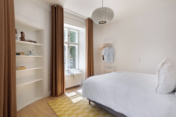 A bedroom with a bed, a window with curtains, a radiator, and a shelf with books and a vase. - Former Dukes Residence - 4 bedrooms - 2 bathrooms (Copenhagen)