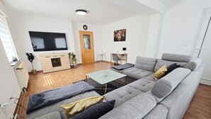 A modern living room with a grey sofa, a small table, and a TV. There is a wooden door and a window with blinds.