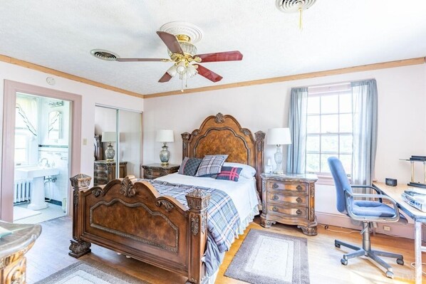 Un dormitorio con cama de madera, un escritorio con silla, un ventilador de techo y una ventana con cortinas.