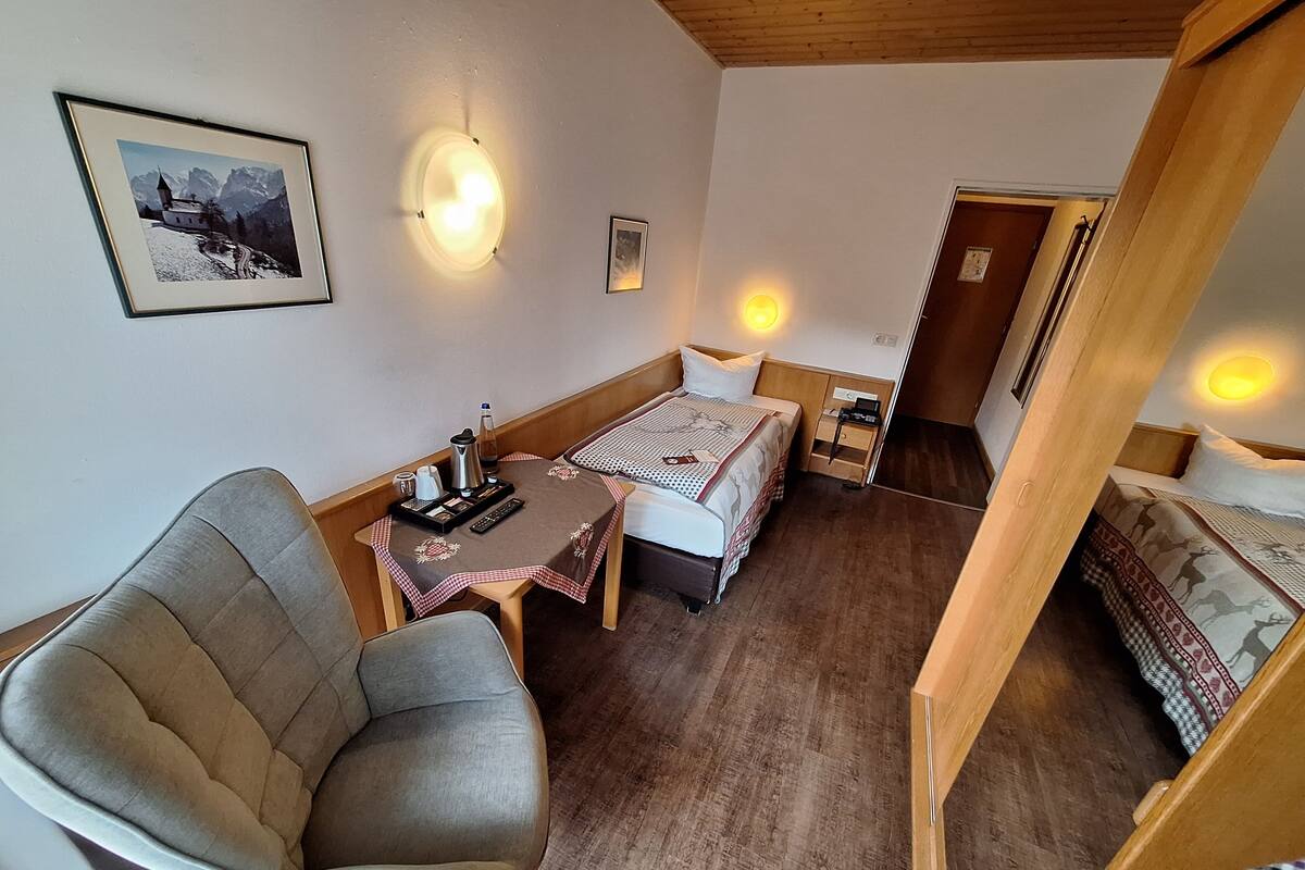 A cozy room with a bed, a chair, a small table with a kettle and cups, and framed pictures on the wall.