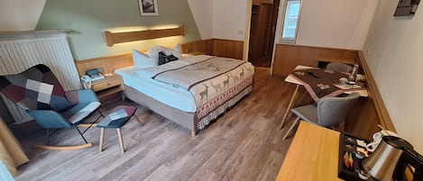A bedroom with a bed, bedside table, chair, desk, and a kitchenette area.