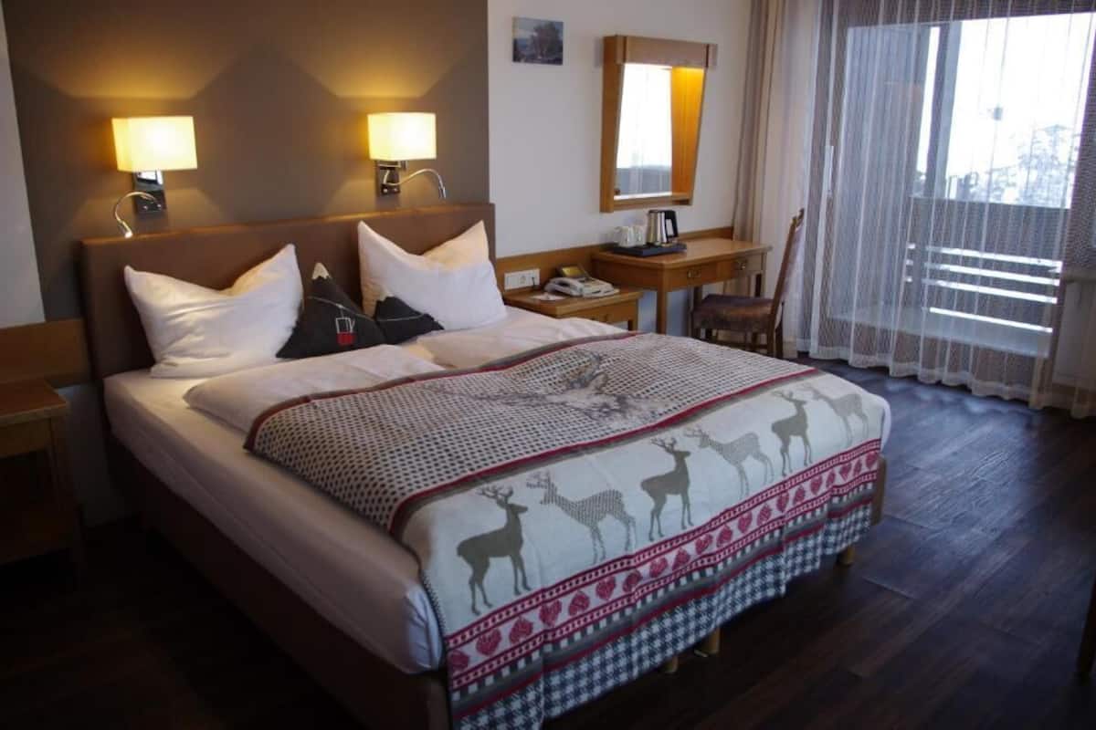 A hotel room with a large bed, two bedside lamps, a nightstand, a chair, and a window with sheer curtains.