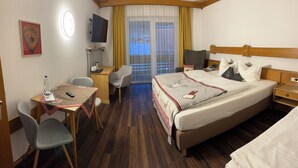 A hotel room with a bed, a desk with a chair, a television, a window with curtains, and a small table with a water bottle and a remote control. - Hotel Löwen (Reit im Winkl)