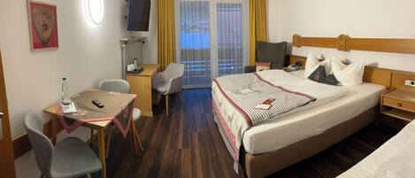 Quarto de hotel com cama, escrivaninha com cadeira, televisão, janela com cortinas e pequena mesa com garrafa de água e controle remoto.