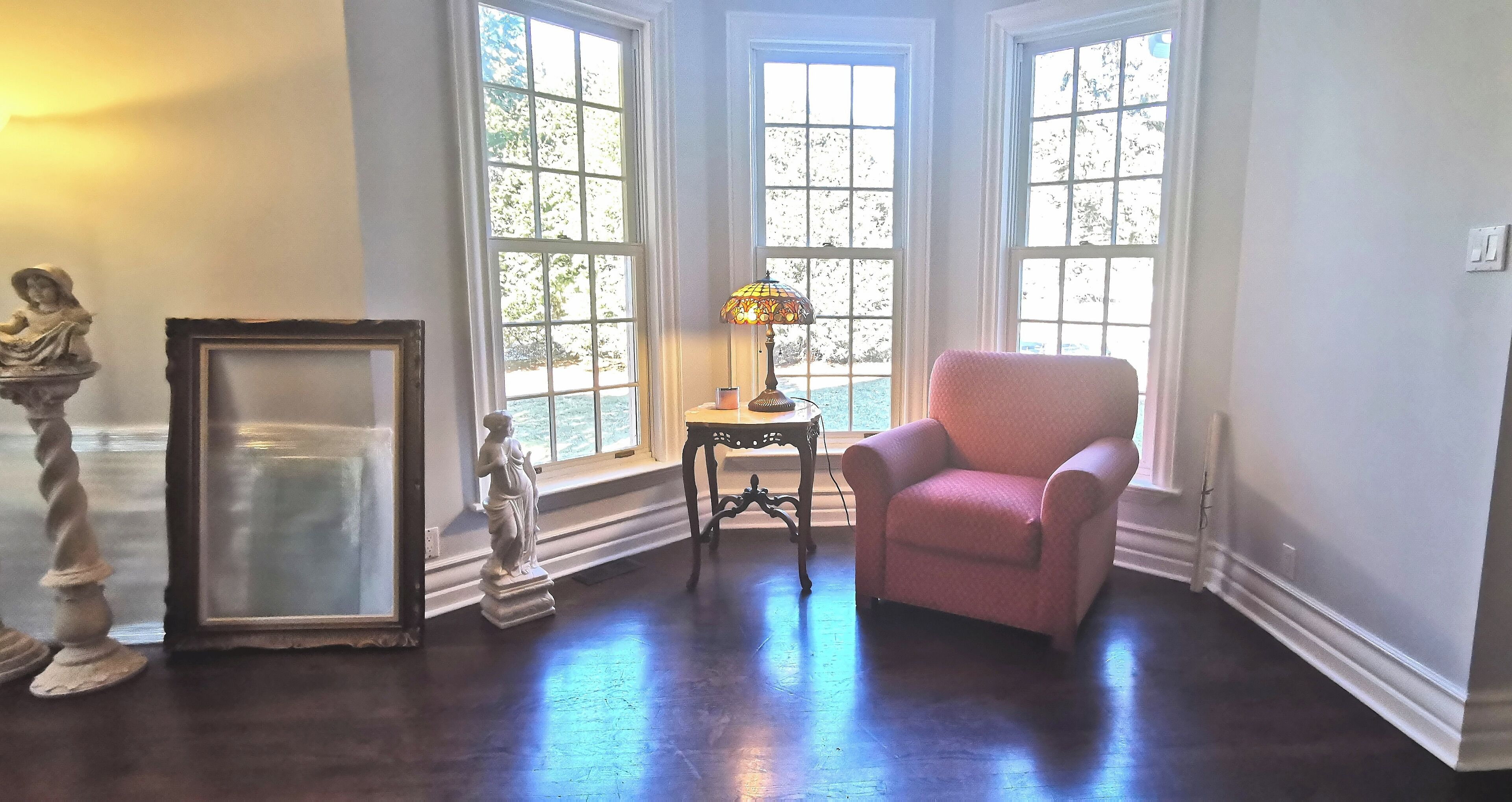 A room with a pink armchair, a statue, a lamp, and a framed picture near a bay window with a view of trees.