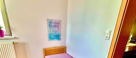 A small, single-bed room with a wooden floor, a radiator, a wall-mounted poster, and a door leading to another room.