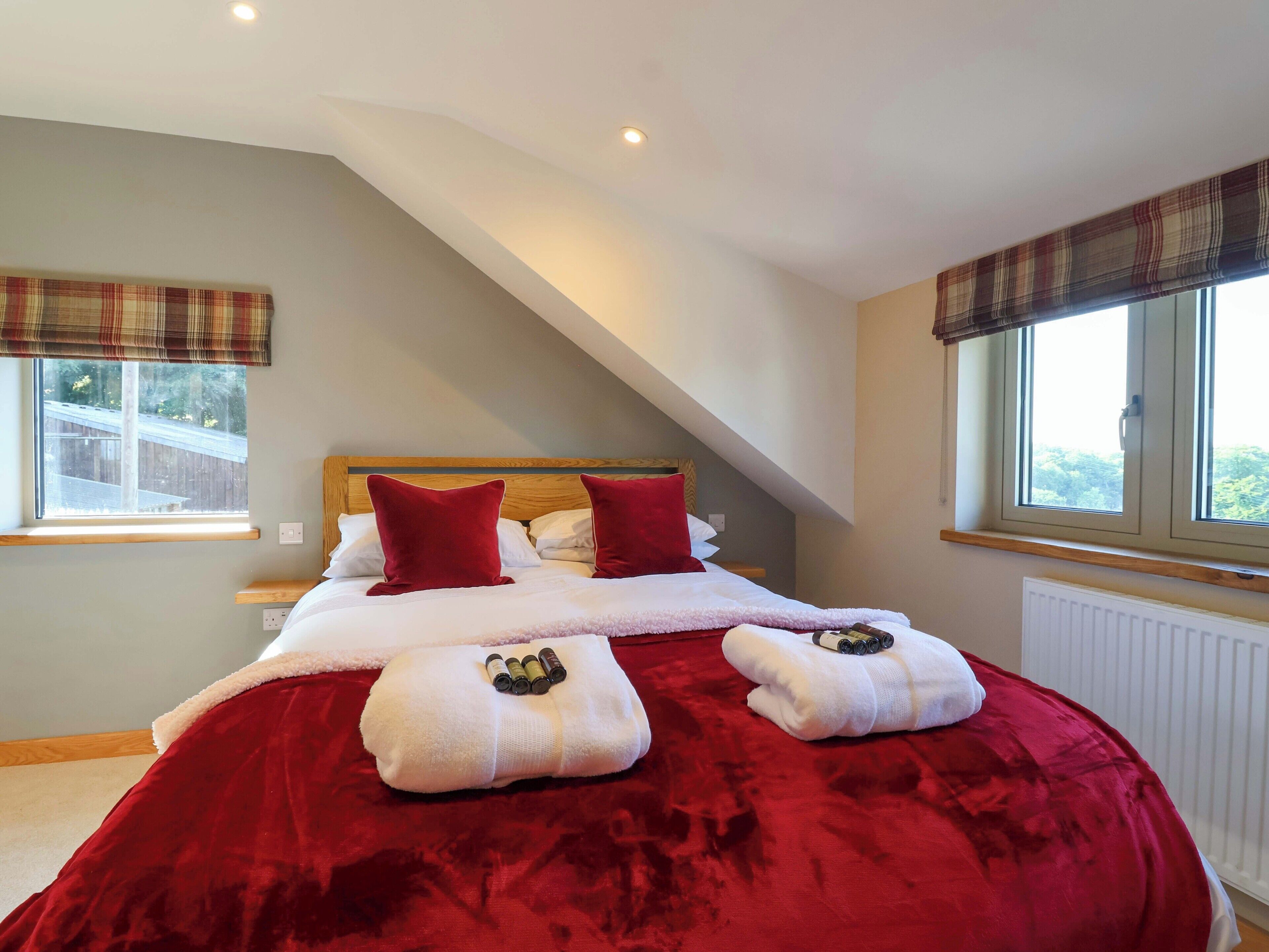 A bedroom with a large bed, red bedding, two pillows, a wooden headboard, and two windows with curtains.