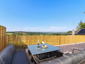 A patio with a table set for two, a wooden fence, and a scenic view of rolling hills. - Heathmoor (Yelverton)